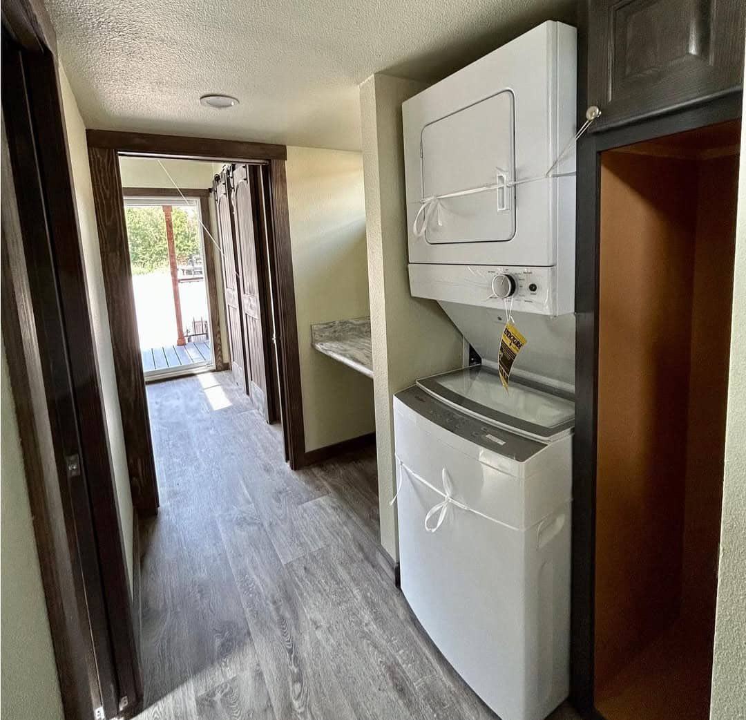 A narrow hallway with gray wood flooring features a white stacked washer and dryer on the right. Sunlight shines through a glass door at the hallway's end.