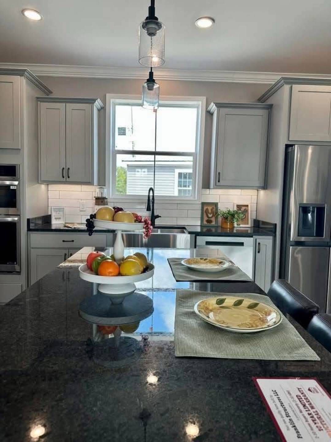 Modern kitchen with gray cabinets, white subway tile backsplash, and stainless steel appliances. A dark granite island holds fruit and plates, under a pendant light.