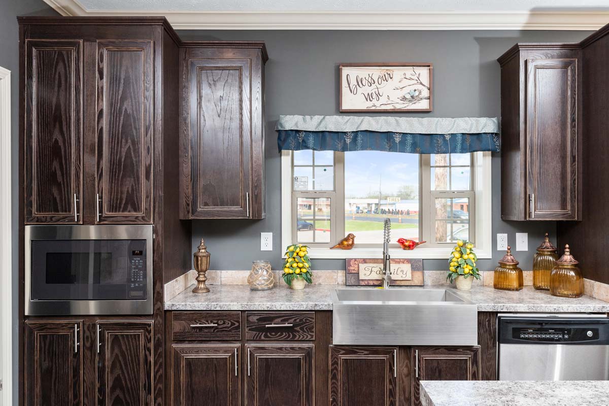 Dark wood kitchen with granite countertops, stainless steel sink, and appliances. A window framed by blue curtains overlooks greenery, adorned with potted flowers.