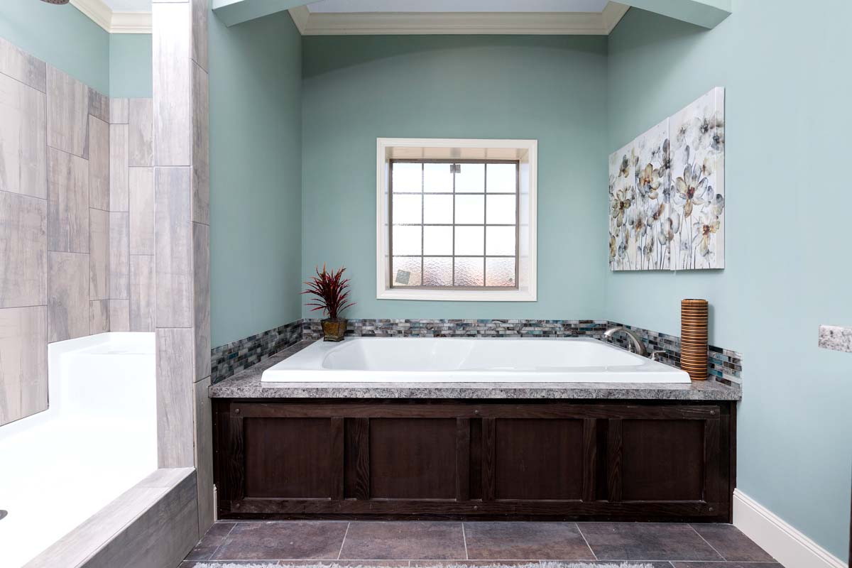 Spacious bathroom with a large white bathtub set against teal walls. Features a window, floral artwork, and decorative items, creating a serene ambiance.