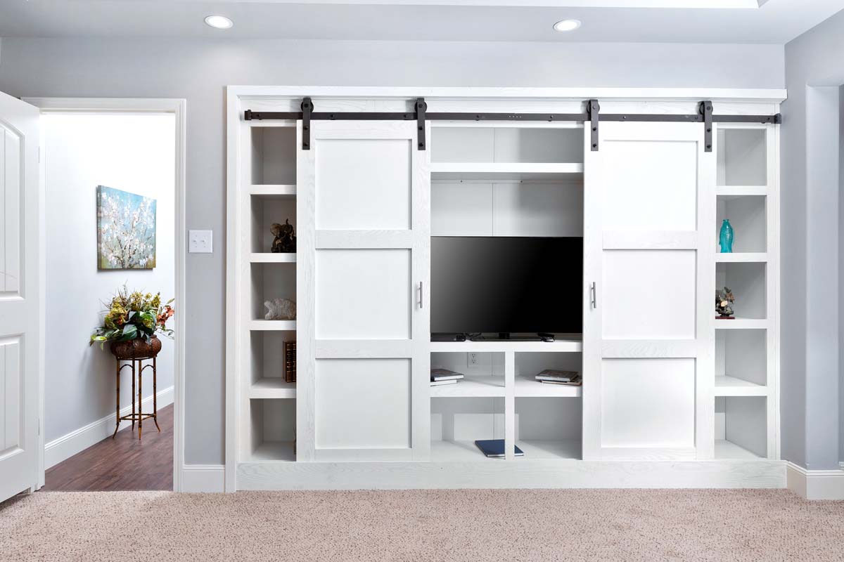 Built-in entertainment unit with white sliding barn doors, revealing a TV and shelves. Cozy room with carpeted floor, open door, and wall art.