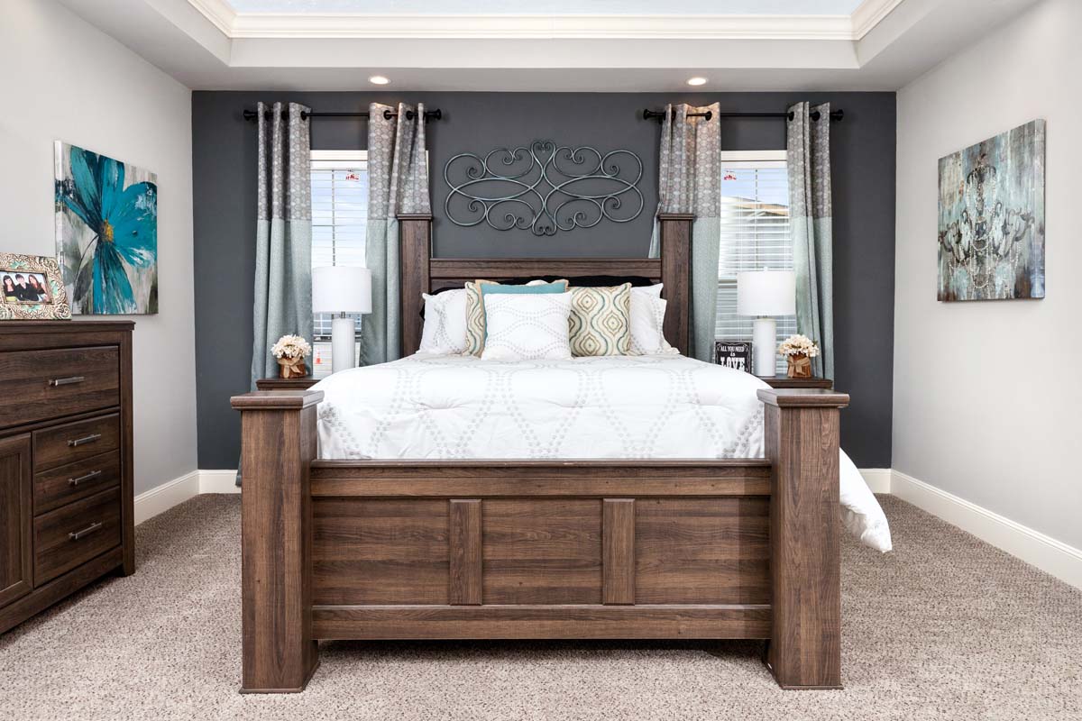 A cozy bedroom with a dark wood bed, white bedding, and patterned pillows. Two teal-curtained windows and matching lamps flank the bed. Walls feature floral paintings, creating a serene ambiance.