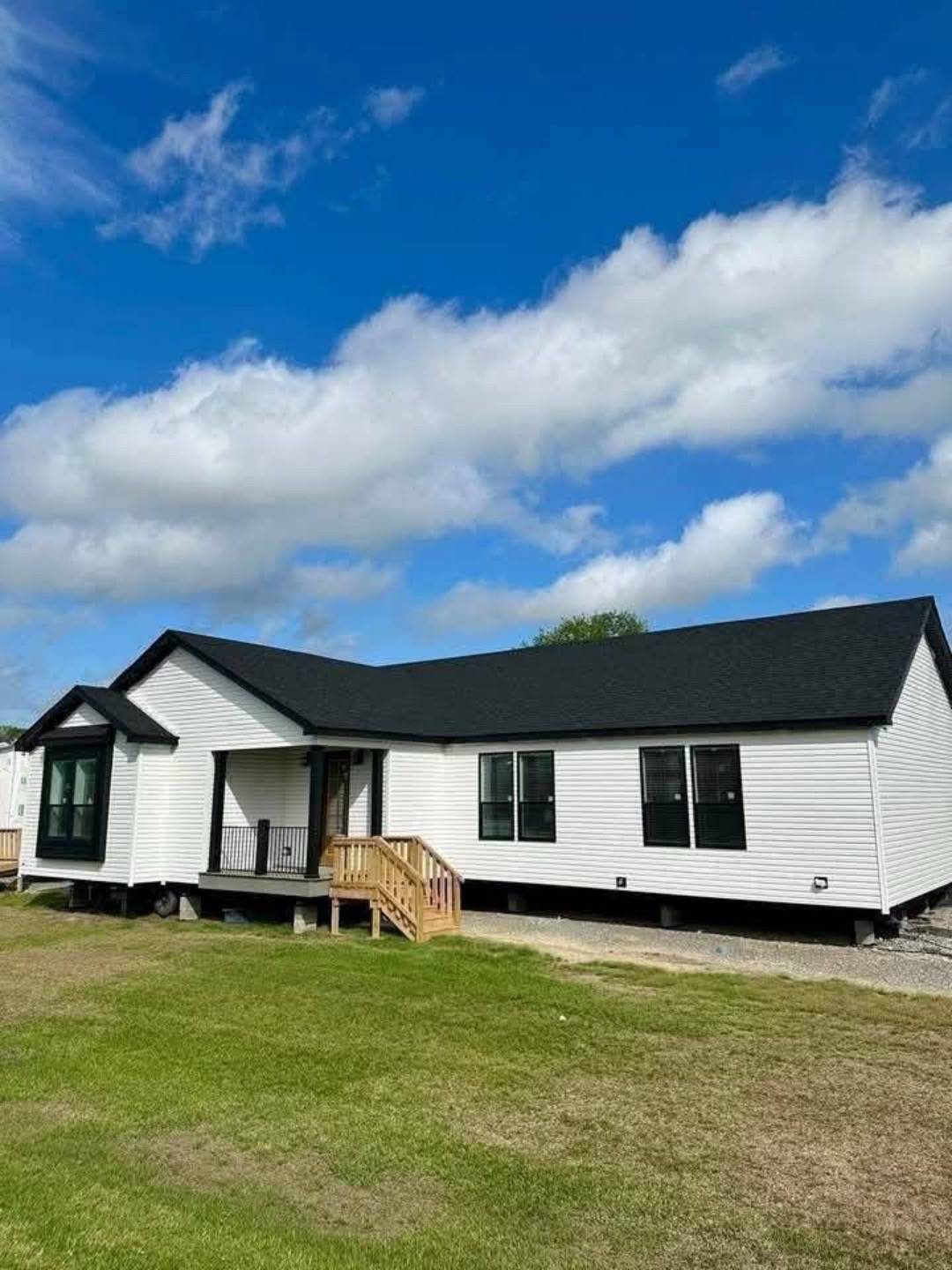 Single-story white house with black roof and covered porch against a vibrant blue sky with fluffy clouds. Surrounded by green grass, creating a peaceful tone.