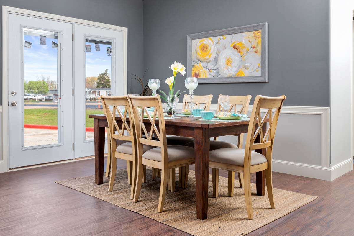 A dining room with a wooden table set for six, teal dishes, and yellow flowers in a vase. Grey walls feature a flower painting. French doors open to a green lawn.