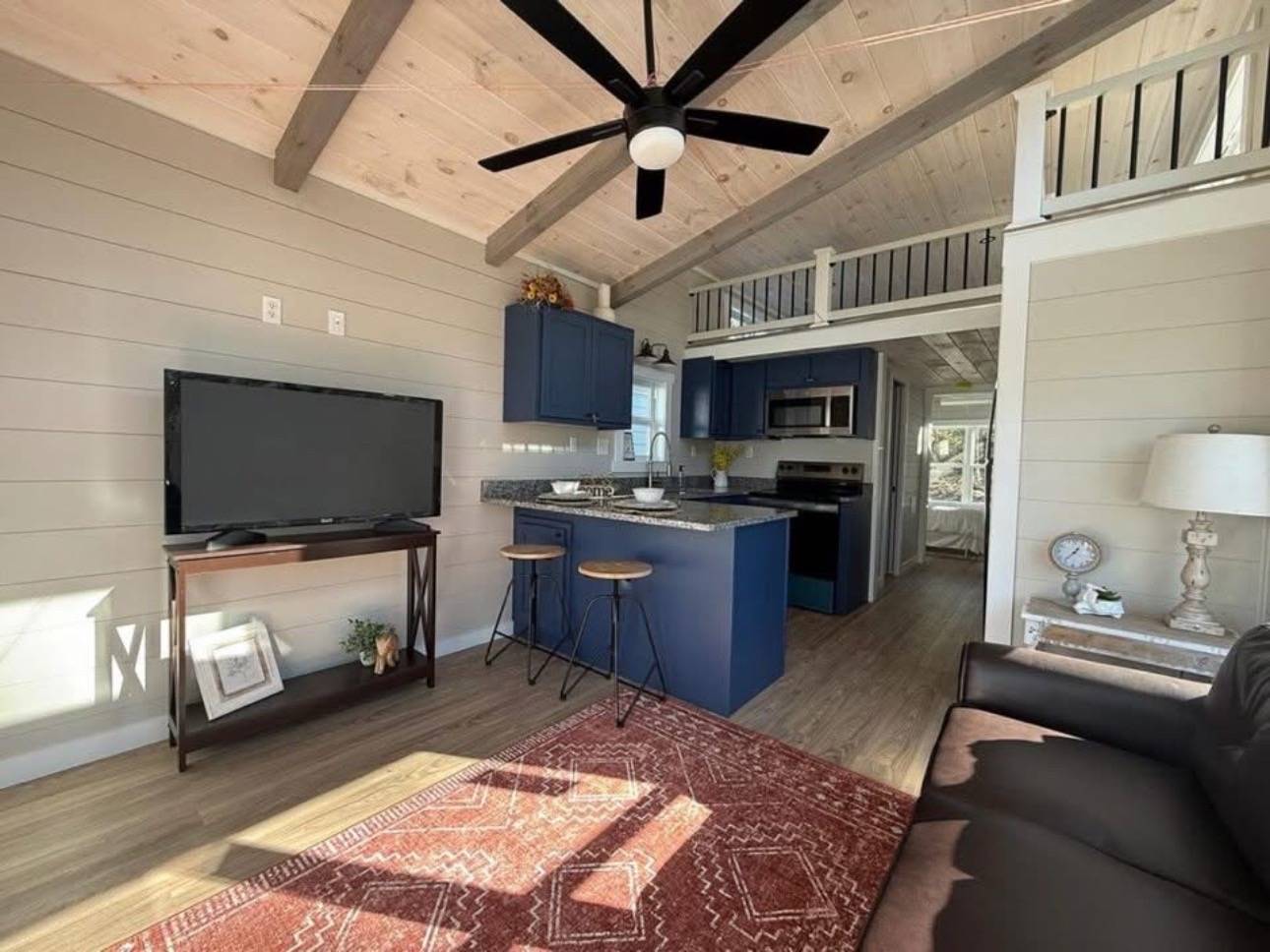 Cozy living area with vaulted ceiling and wooden beams. Features include a dark fan, navy cabinets, island with stools, TV, and red rug, conveying warmth.