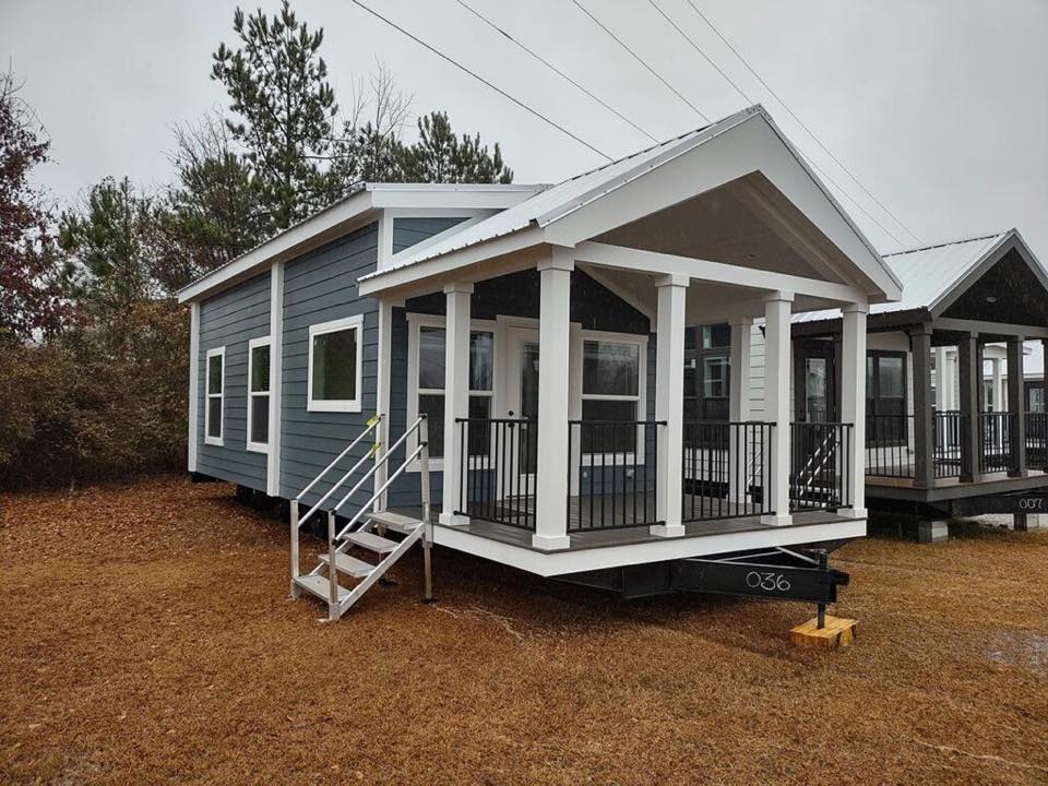 A small, modern tiny house with blue siding and white trim sits on a grassy lot. It features a covered porch with stairs, surrounded by trees under an overcast sky.