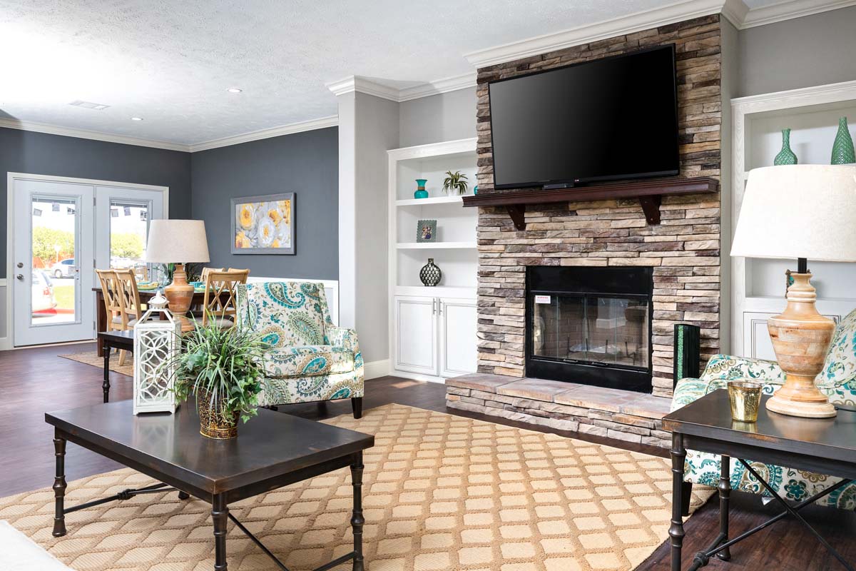 Stylish living room with a stone fireplace and mounted TV. Features patterned chairs, wooden tables, and a beige rug. Light and airy ambiance.