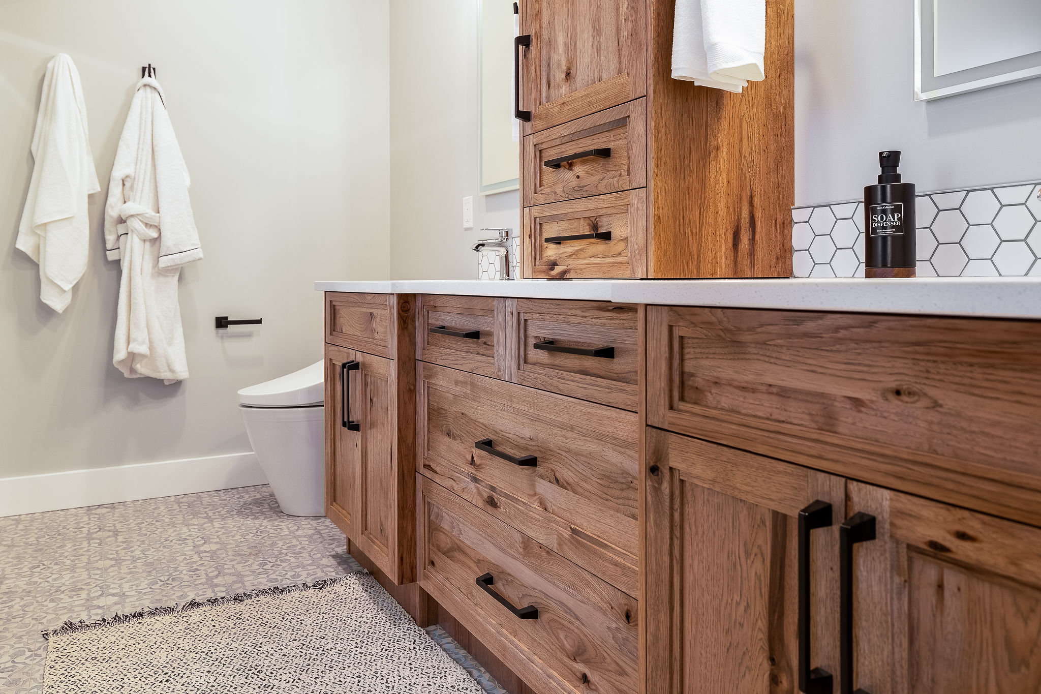 Rustic bathroom with wooden cabinets and black handles, featuring a white countertop, geometric tiles, two white robes, and a modern toilet.