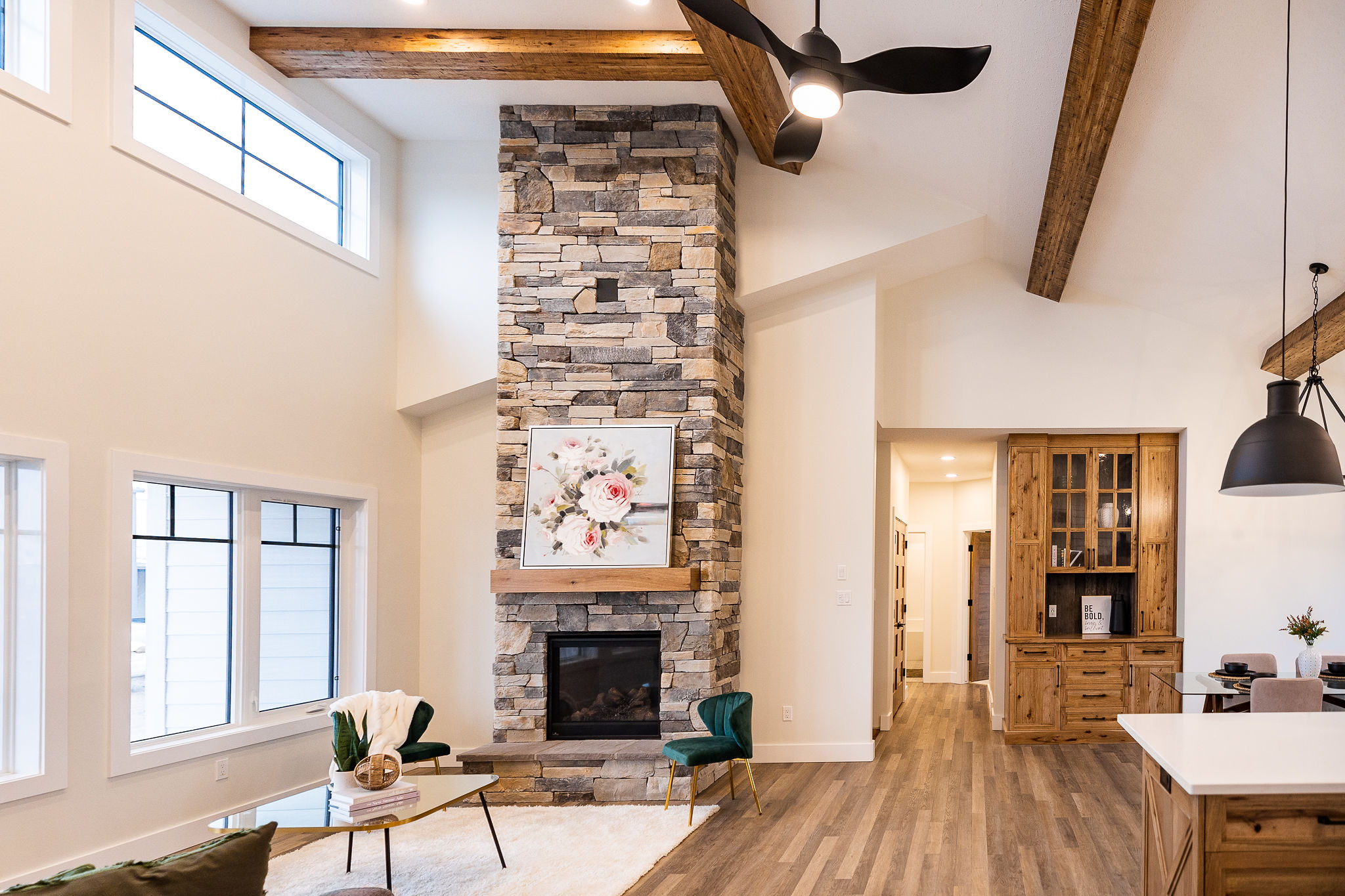Spacious living room with a stone fireplace, vaulted ceiling, and wooden beams. Elegant decor includes a floral painting and modern furniture. Cozy and inviting.