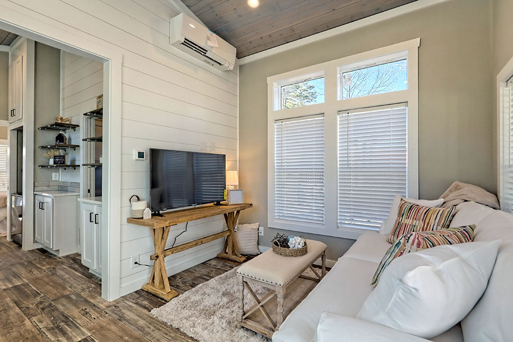 Cozy living room with a white sofa, colorful cushions, and a rustic wooden bench. A TV on a wooden console, large windows, and soft lighting create a warm atmosphere.