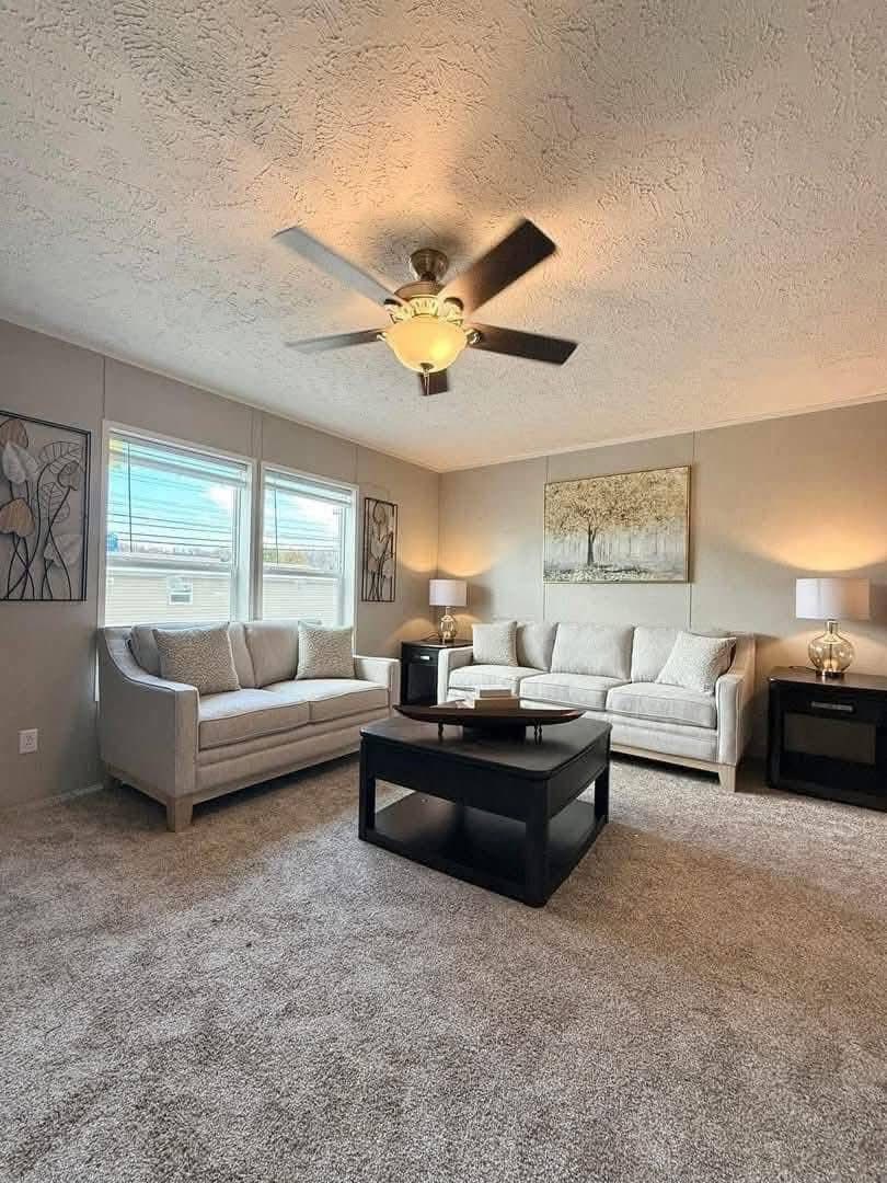 Spacious living room with neutral tones featuring two cream sofas, a black coffee table, abstract wall art, lamps, and a ceiling fan, creating a warm, inviting atmosphere.