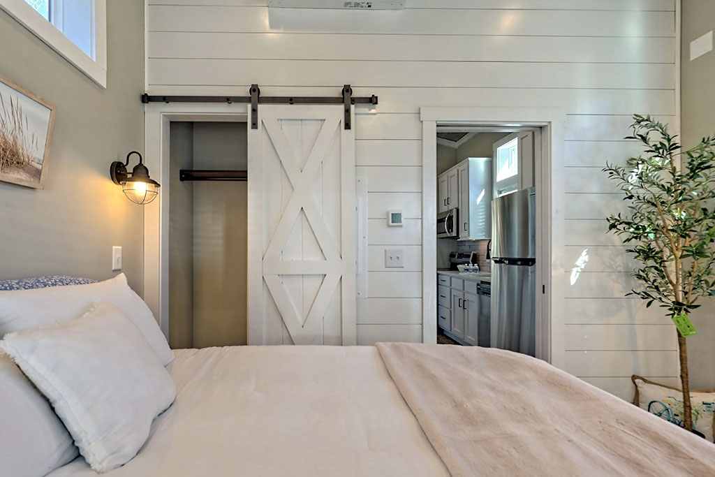 A cozy bedroom with white shiplap walls features a barn-style door leading to a closet. A soft bed, sconce lighting, and a glimpse of a kitchen enhance warmth.