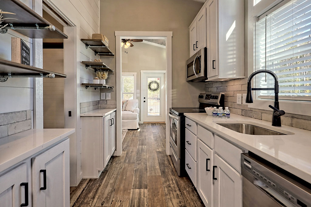 Modern kitchen with white cabinets, stainless steel appliances, and a farmhouse sink. Wooden floor leads to a bright living room with a door and wreath.
