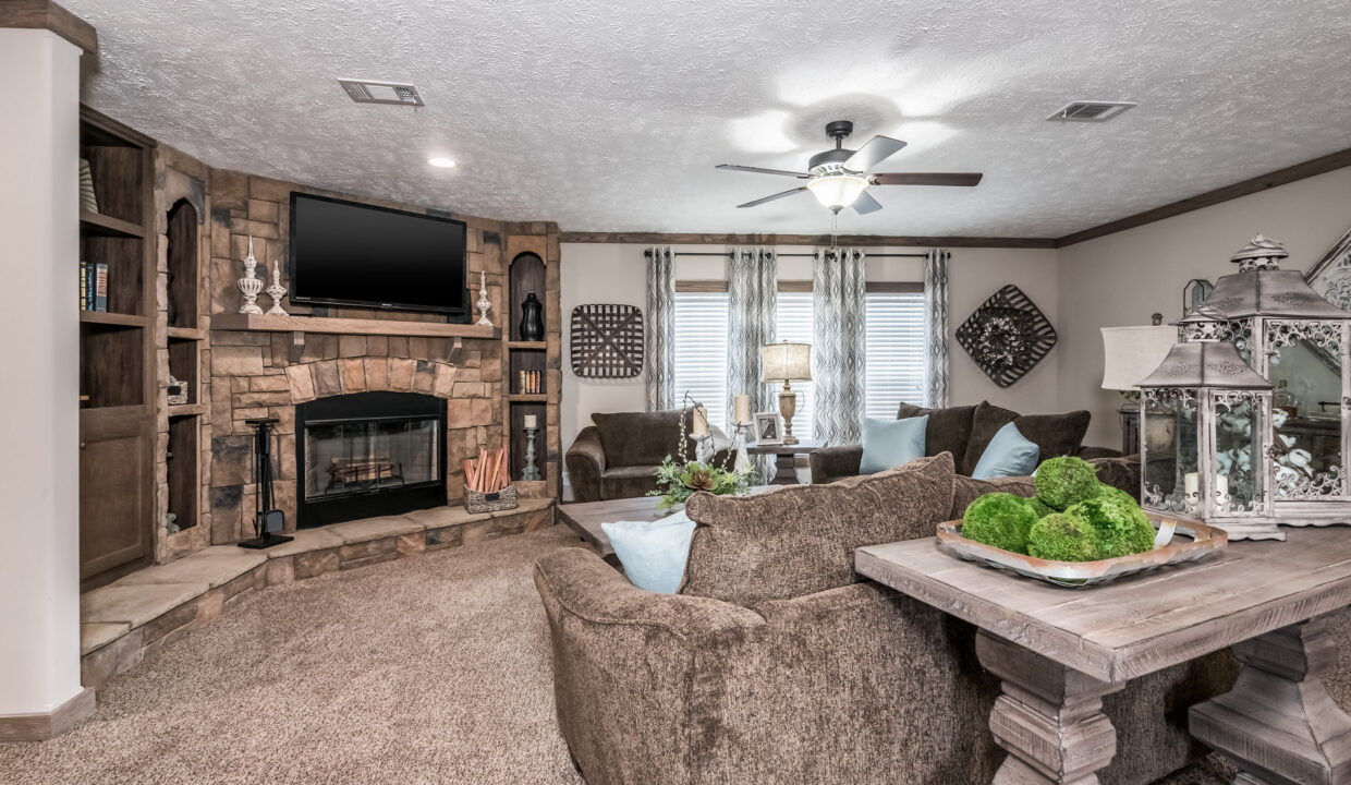Cozy living room with brown sofas, stone fireplace, and TV. Decorative plants on a wooden table, ceiling fan, and natural light through large windows.