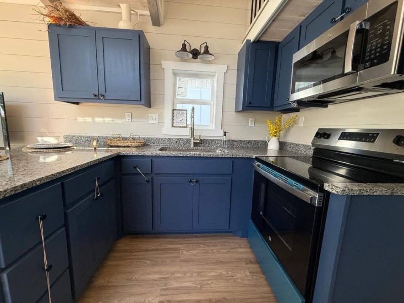 Cozy kitchen with navy blue cabinets, granite countertops, and stainless steel appliances. Wooden floor, white window, and rustic decor add warmth.