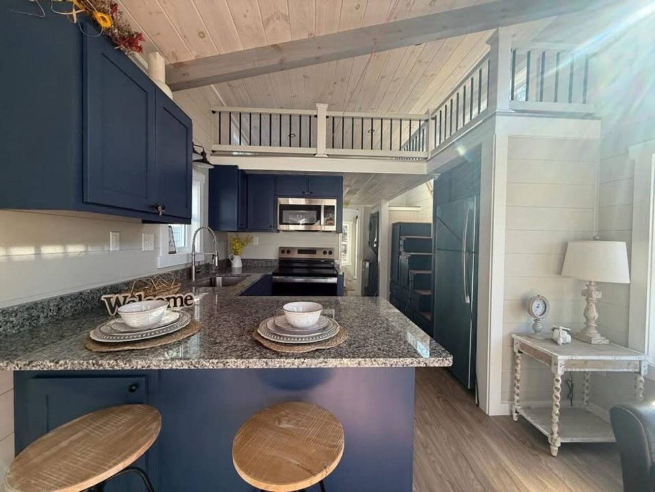 Cozy kitchen with blue cabinets, granite countertops, and a loft above. Barstools at the counter, warm sunlight, rustic charm, and welcoming ambiance.