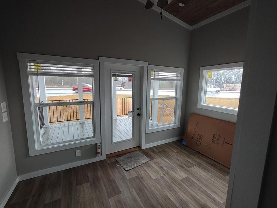 A small, bright room with three large windows and a glass door leading to a porch. The wood floor and gray walls create a cozy, modern feel.