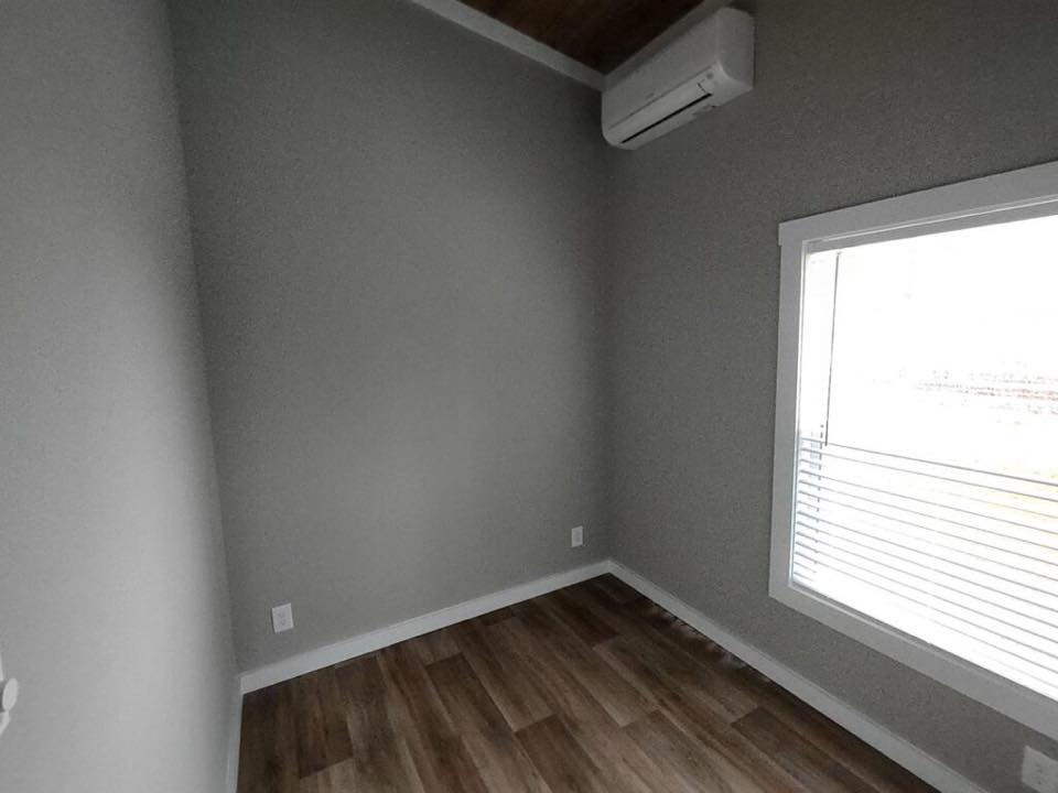 Small room with gray walls and wooden flooring. A large window with blinds lets in light. An air conditioning unit is mounted high on the wall.