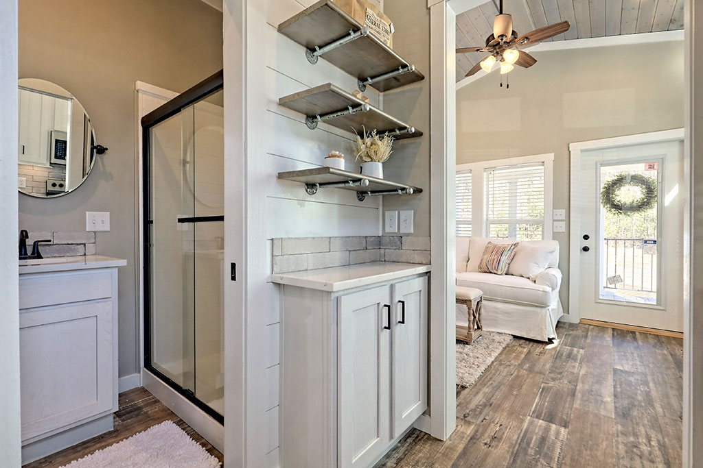Stylish modern interior; view of a cozy living room with white sofa and ceiling fan, adjacent to a compact bathroom with glass shower and round mirror.