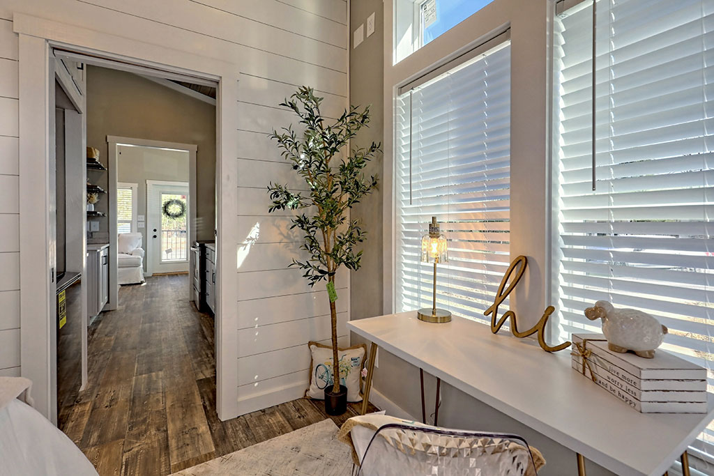 Bright home office with cozy decor and natural light pouring through large windows. Features a white desk, a decorative lamp, and an indoor plant.