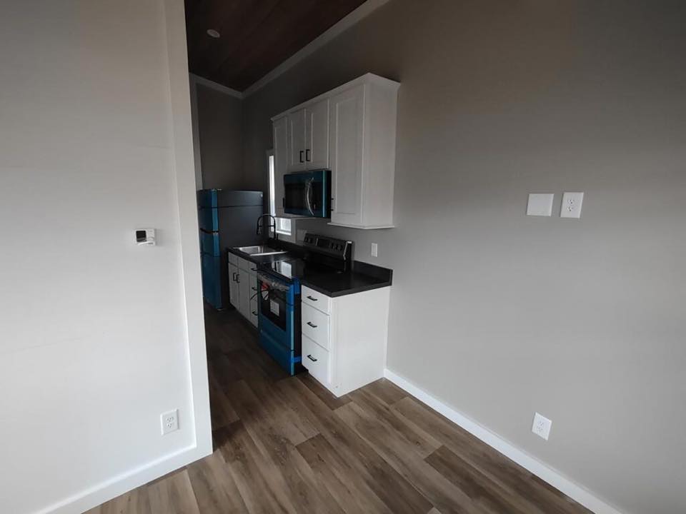 Compact kitchen with white cabinets, black countertops, and a blue-accented stove. Wooden floor and gray walls create a modern, clean look.