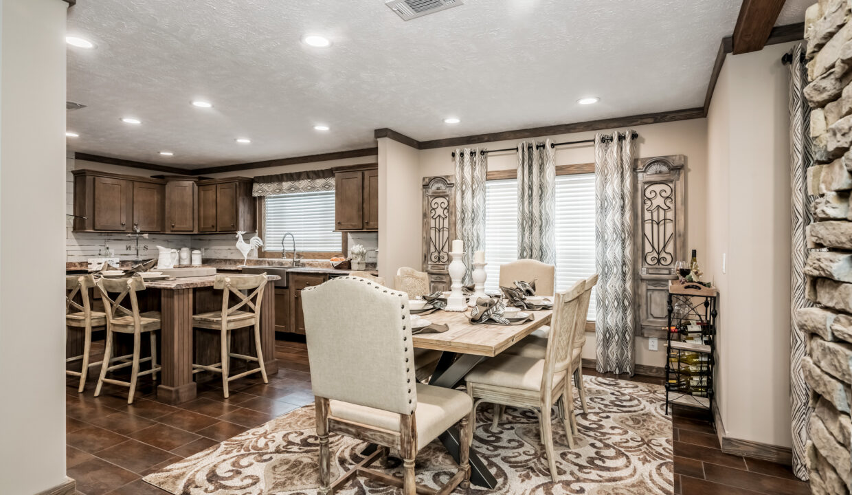 A cozy dining room with wooden furniture and earth-toned decor. A dining table is set for six, adjacent to a rustic kitchen with wooden cabinets. Soft lighting creates a warm, inviting atmosphere.