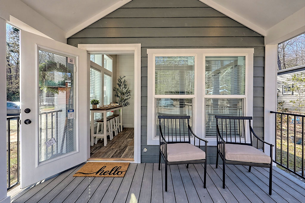 A cozy porch with two black chairs and beige cushions on gray wood flooring. Open glass door showing dining area inside. A doormat reads "hello."