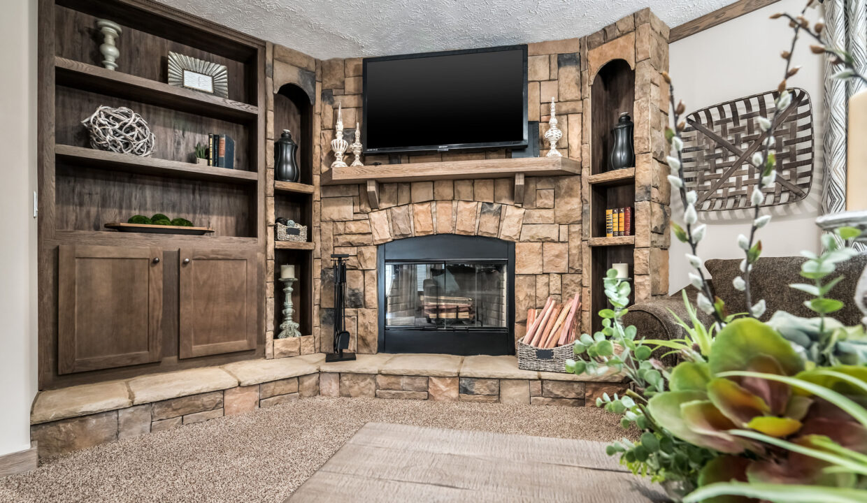 A cozy living room with a stone fireplace, flanked by wooden shelves and a TV above. Decor includes candles, books, plants, and wicker accents.