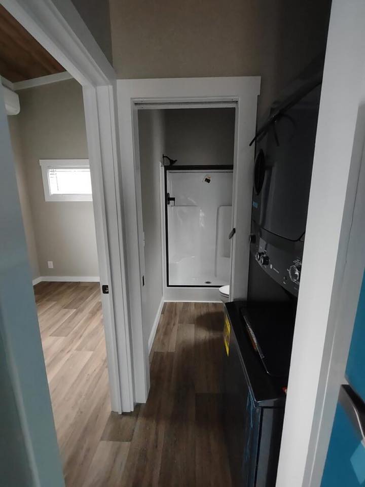 Narrow hallway with wood flooring leads to a compact bathroom. A small shower is on the right, and a stacked washer-dryer is visible on the left. Bright, tidy, and functional.