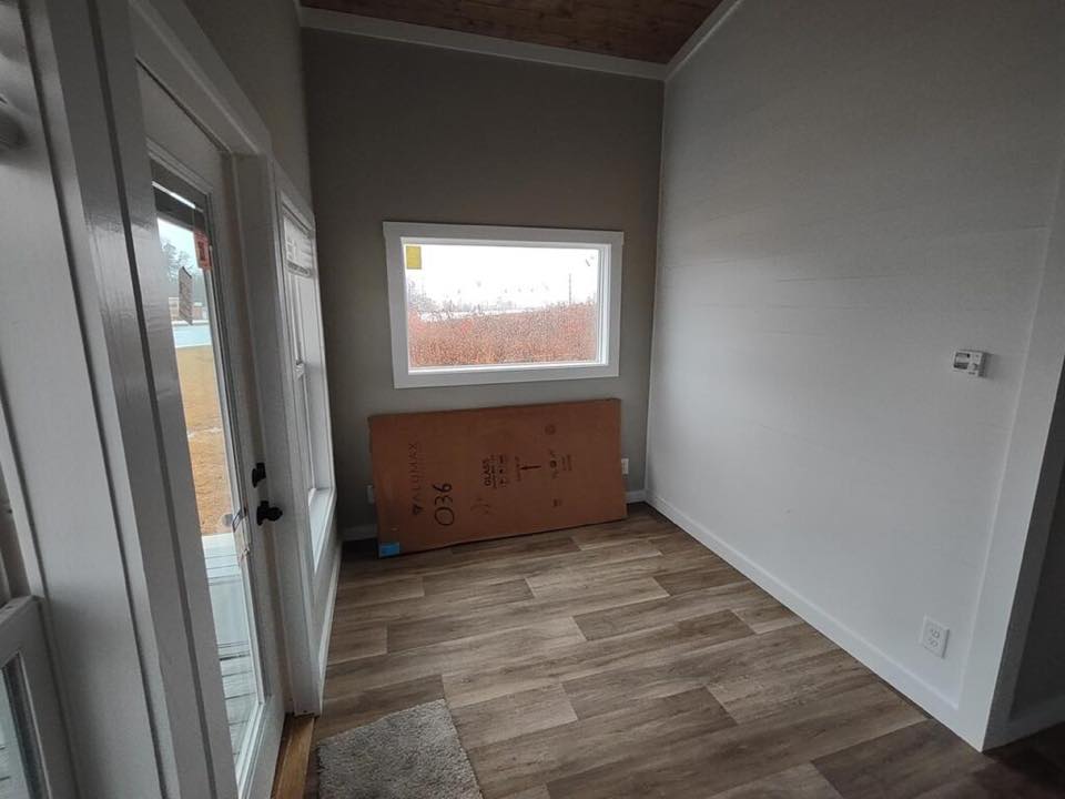 A small, empty room with light wood floors and gray walls features a large window. A cardboard box leans against the wall, and a glass door opens to the outside.