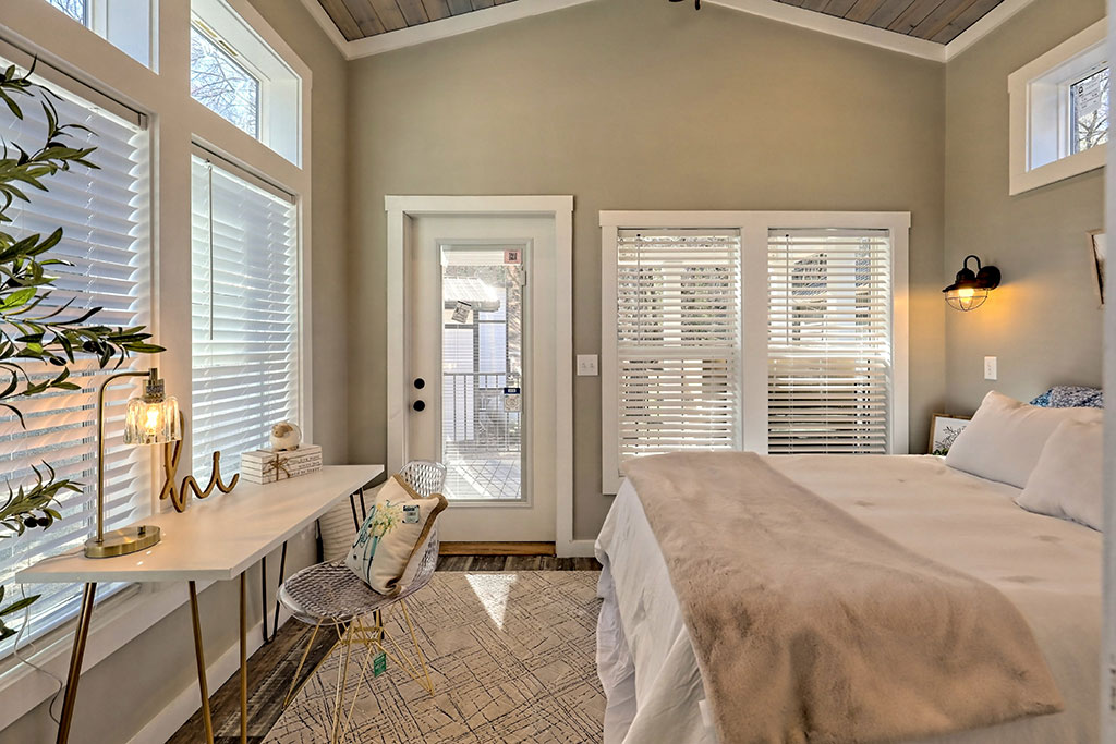 Bright and cozy bedroom featuring a large bed with a beige blanket, a modern desk with a lamp and decor, tall windows with blinds, and a glass door.