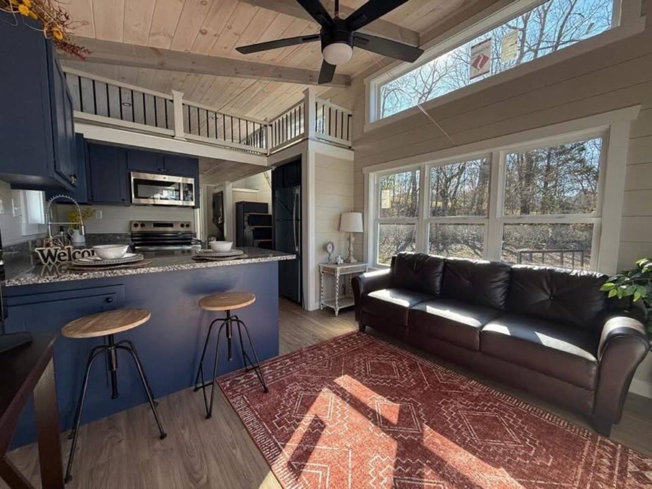 Cozy cabin interior with vaulted ceiling, featuring a blue kitchen, bar stools, leather couch, and large windows. Sunlit red rug adds warmth.