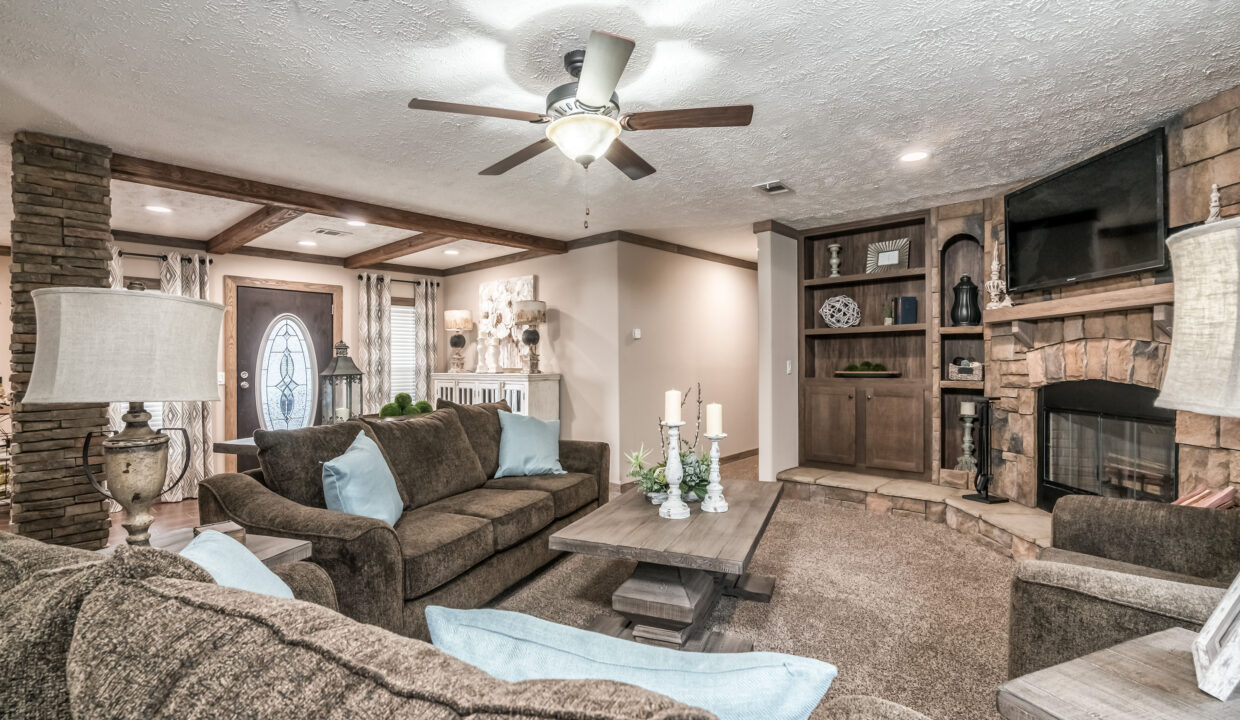 A cozy living room with brown sofas and light blue cushions arranged around a wooden coffee table. There's a stone fireplace with a TV above it, built-in shelves, and decorative candles. A ceiling fan hangs from the textured ceiling. The room has a warm, inviting atmosphere.