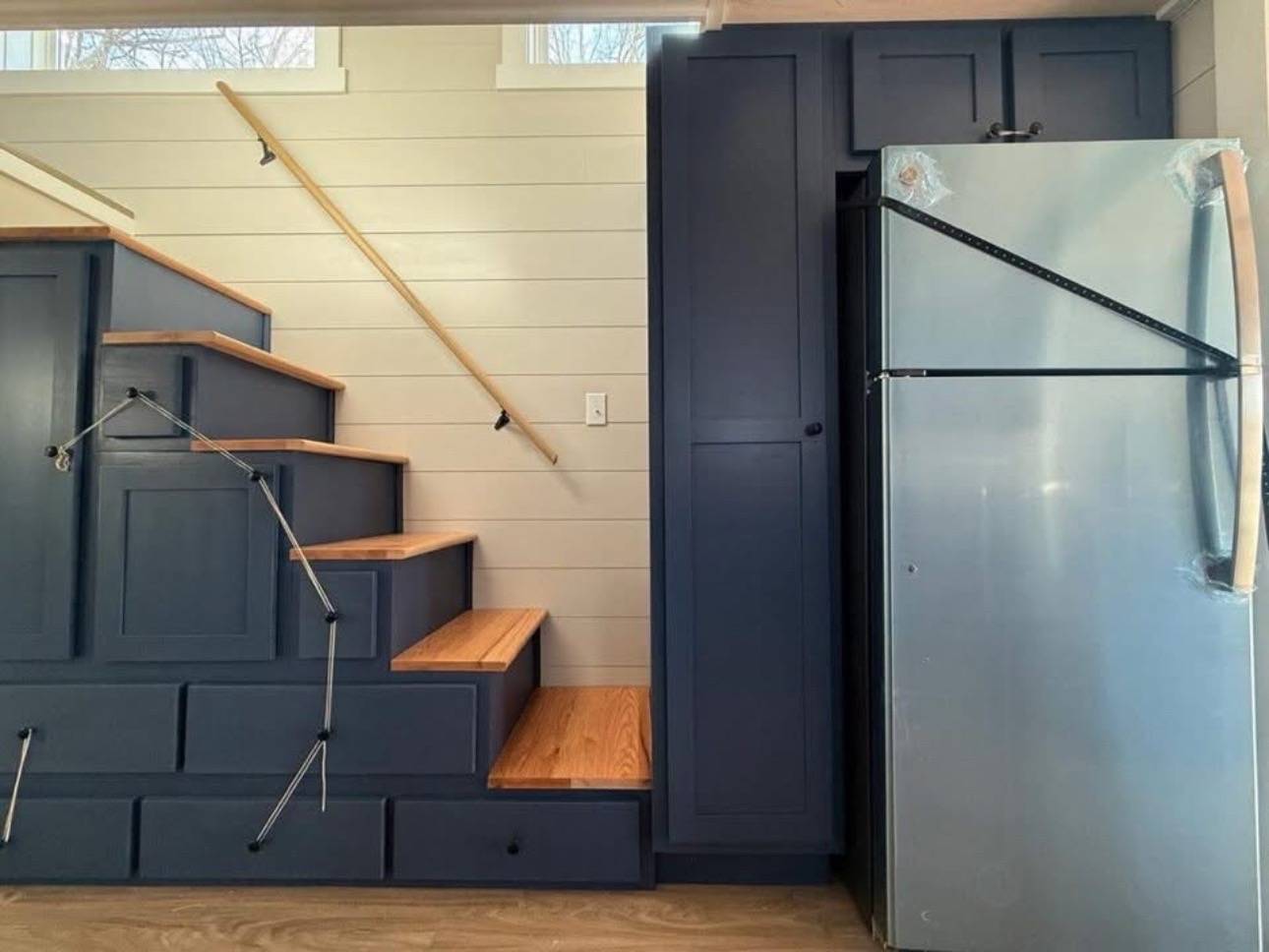 Blue cabinetry staircase with wooden steps and drawers underneath, next to a silver fridge. Light wood-panelled wall with a modern, cozy feel.