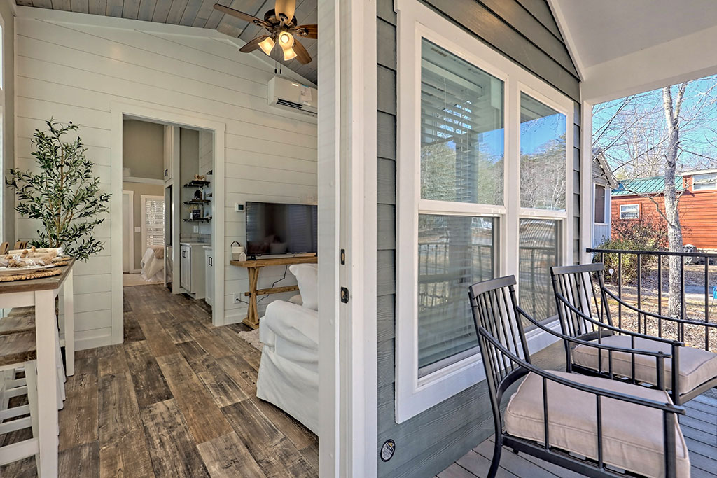 Narrow kitchen with white cabinets, stainless steel appliances, and wooden floor. Open shelves on left, sink and window on right. Sunlit, cozy feel.