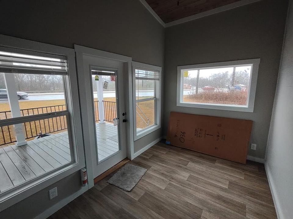 Cozy room with large windows and a glass door leading to a porch. Outside, a wet road, parked cars, and overcast sky are visible, creating a tranquil mood.