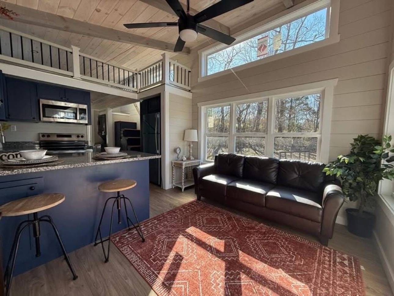 Spacious living room with high ceilings, large windows, and natural light. Features a dark leather sofa, red patterned rug, blue kitchen island, and ceiling fan. Cozy and modern ambiance.