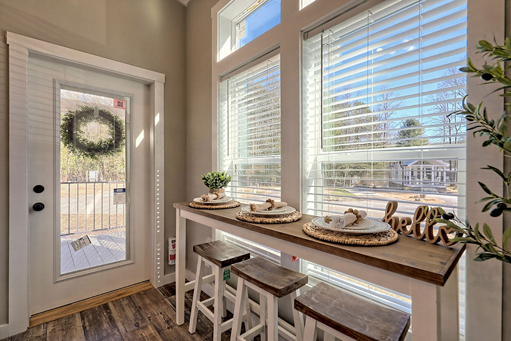 Bright, cozy breakfast nook with three stools, a wooden table with place settings, a small plant, and "hello" decoration. Sunlight streams through large windows.