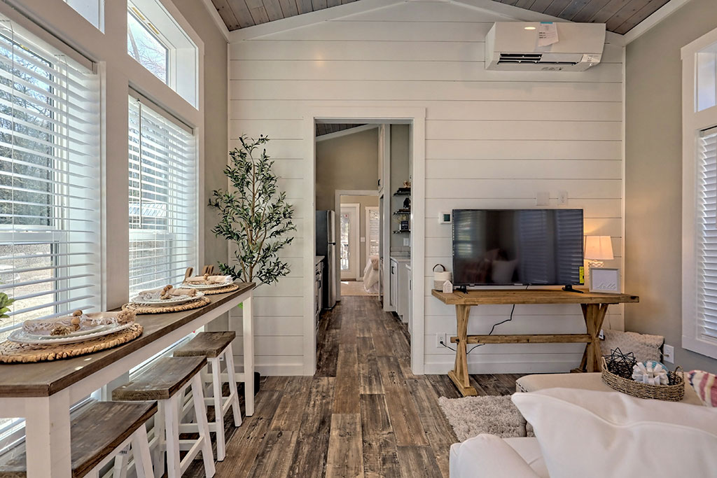 Cozy living area with wooden floors and white shiplap walls. A TV on a wooden stand, large windows with blinds, and a bar table with stools add warmth.