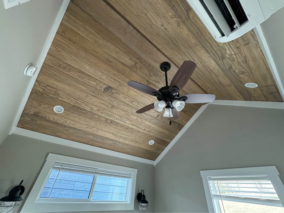 Wooden plank ceiling with a dark ceiling fan centered, bordered by light gray walls. Windows with white trim and soft natural light below.