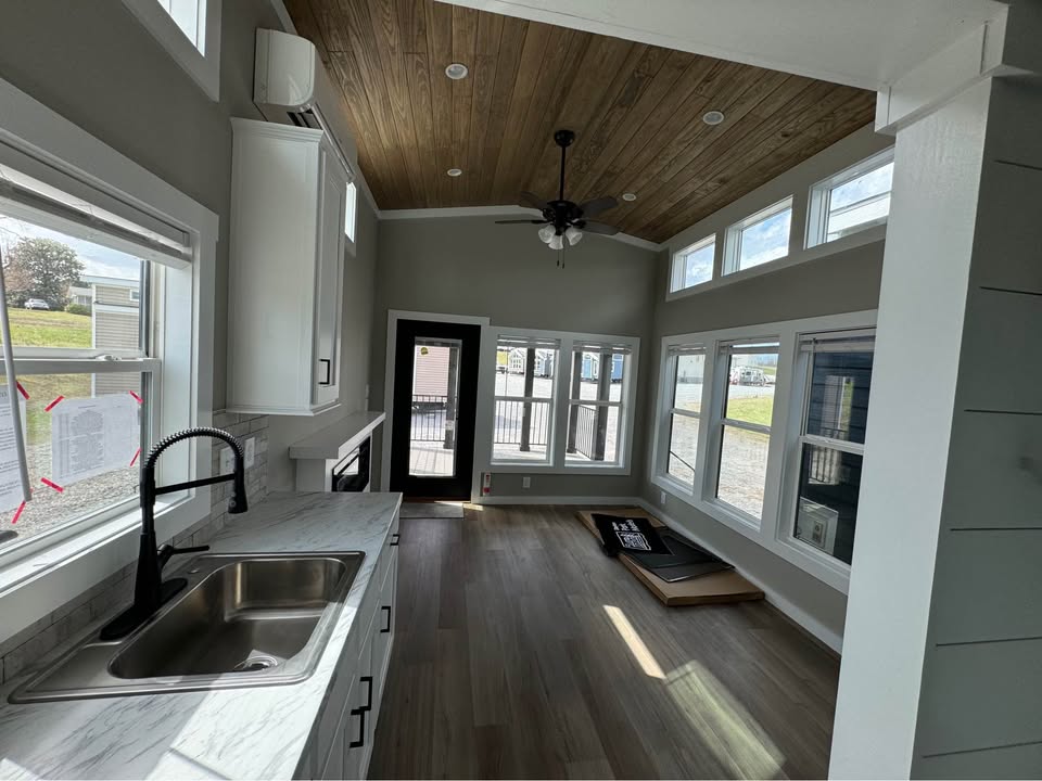 Modern tiny home interior with wood ceiling, gray walls, and large windows. Features a small kitchen with a marble counter, stainless sink, and black faucet. Bright and airy atmosphere.