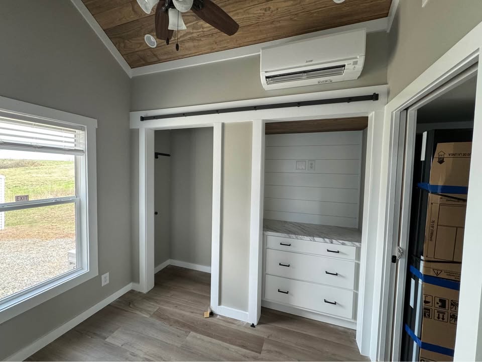 Small room with wood-paneled ceiling and light gray walls. Features a large window, sliding closet doors, and air conditioner. Bright and minimalistic ambiance.