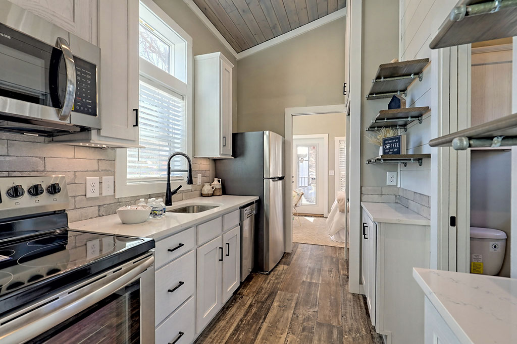 Compact kitchen with stainless steel appliances, white cabinets, and dark wood floors. Open shelves on the right and large window above the sink, creating a cozy, inviting space.
