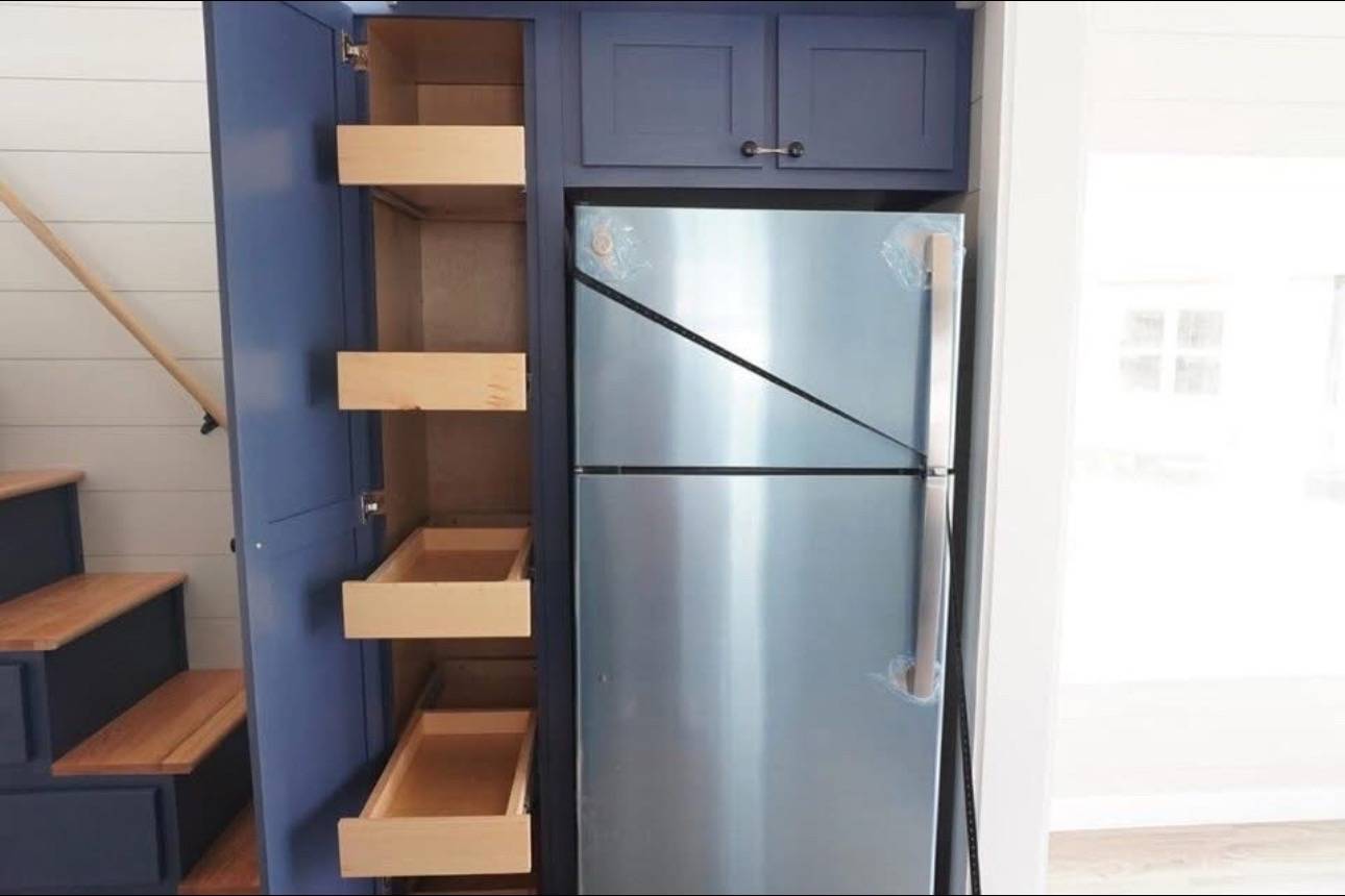 Blue kitchen cabinet with three open wooden pull-out drawers next to a stainless steel fridge. Wooden stairs visible on the left. Bright, modern vibe.
