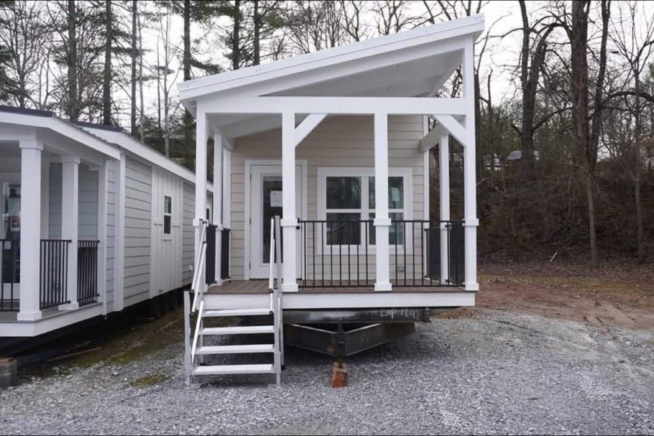 Small modern tiny house with a slanted roof, porch, and ladder-style stairs, set on a gravel area with a wooded background, conveying simplicity.