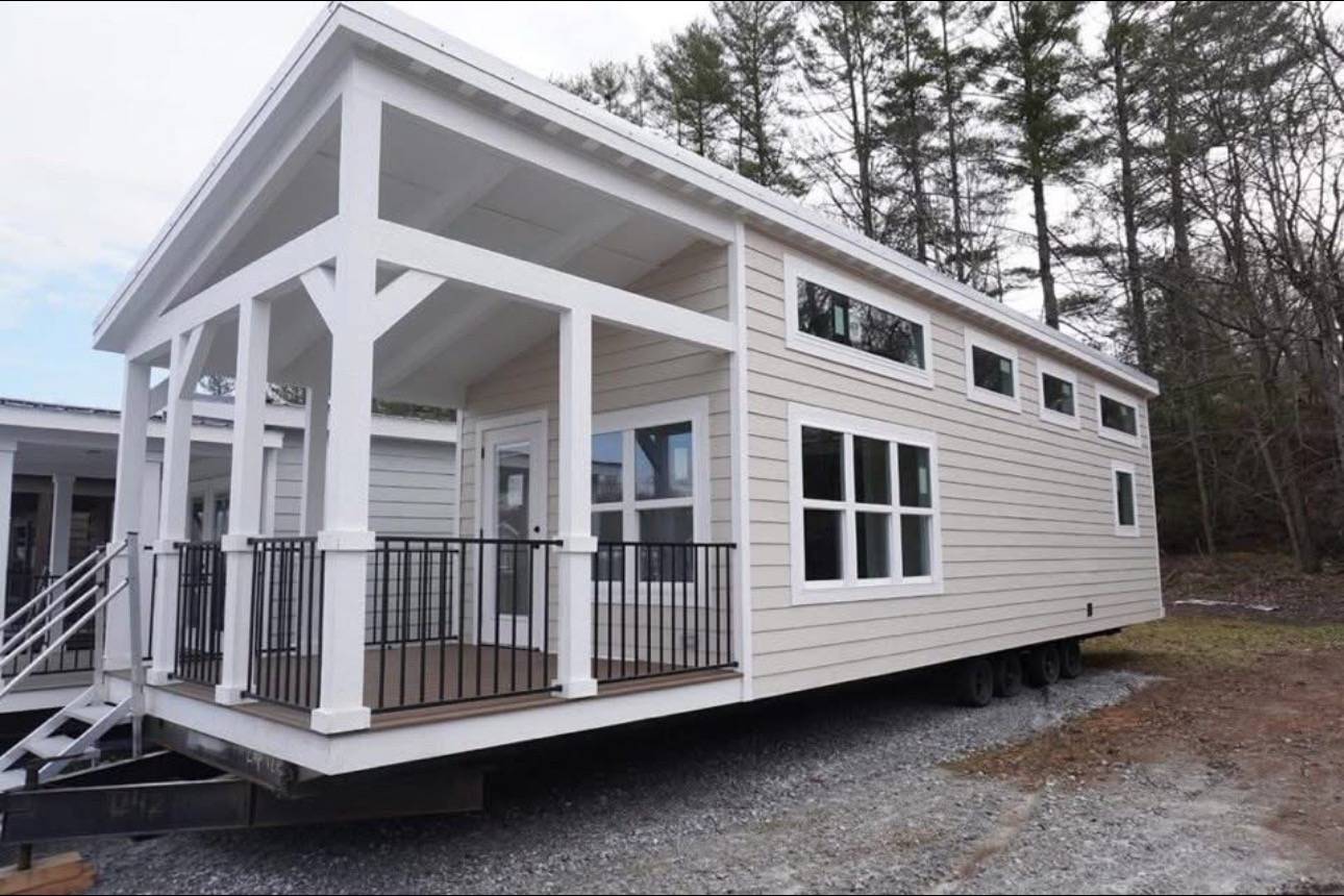 Modern tiny house on a mobile platform, beige siding, black railing, spacious porch, surrounded by trees, conveying a sense of cozy minimalism.