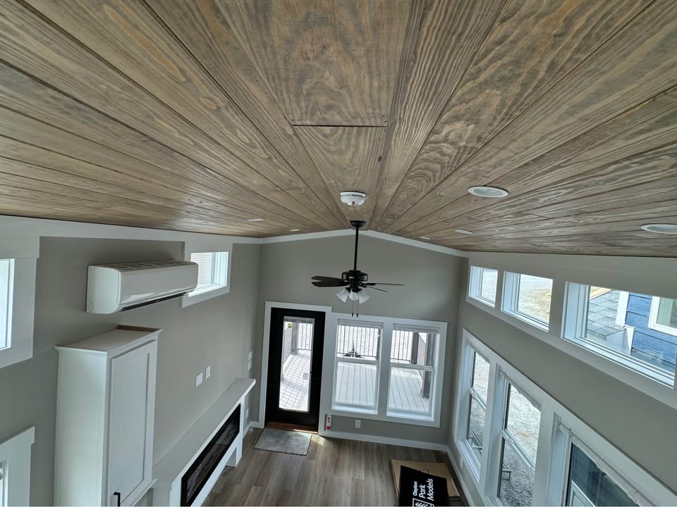 High-angle view of a modern living room featuring a wooden ceiling with a ceiling fan, light grey walls, and large windows. Bright and airy ambiance.
