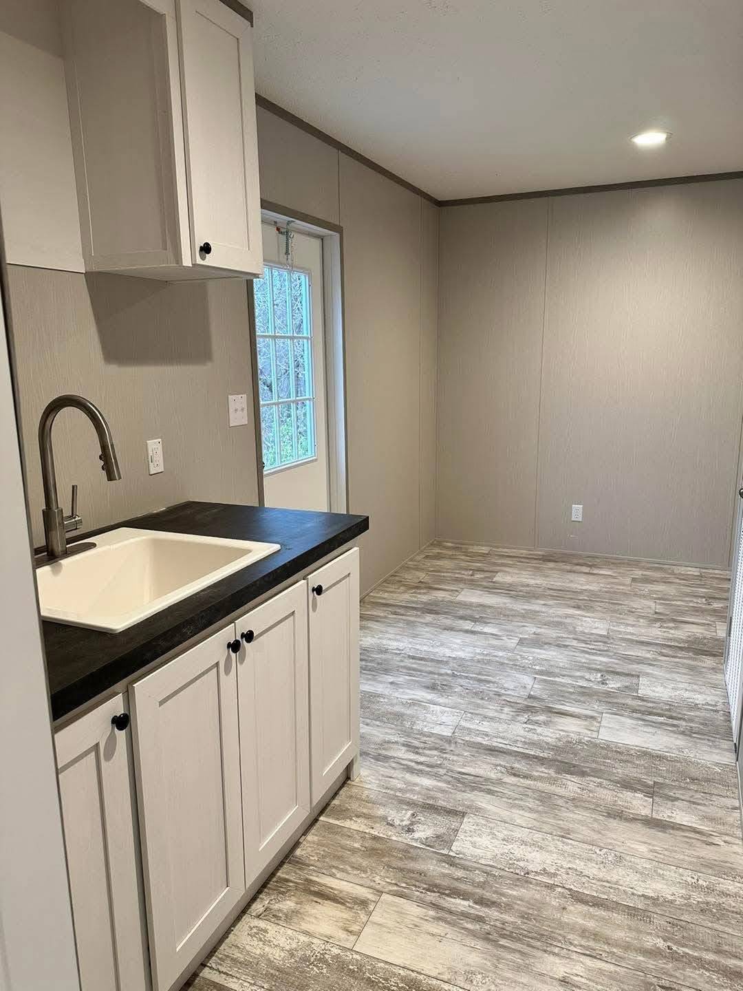 Minimalist kitchen space with white cabinets and a black countertop holding a sink on the left. Gray wooden floors and neutral walls create a calm tone.