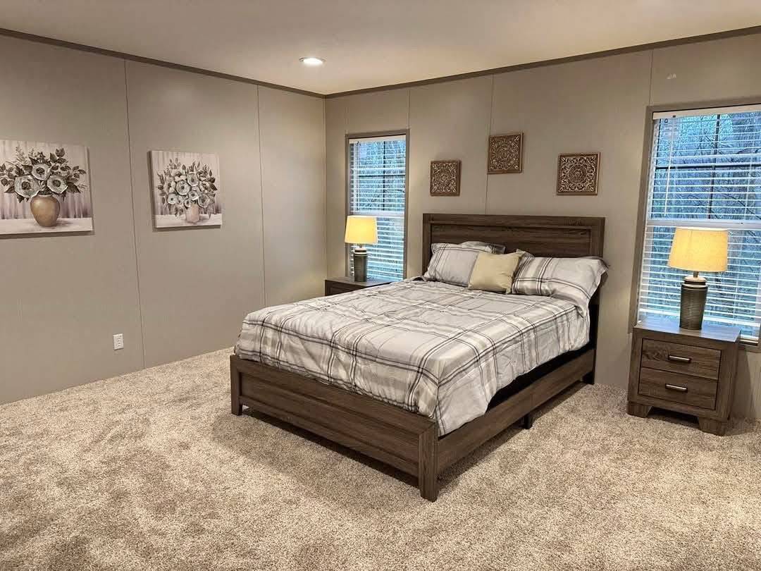 A cozy bedroom with a neatly made plaid bed, flanked by two nightstands with lamps. Soft carpet, floral paintings, and warm lighting create a serene ambiance.