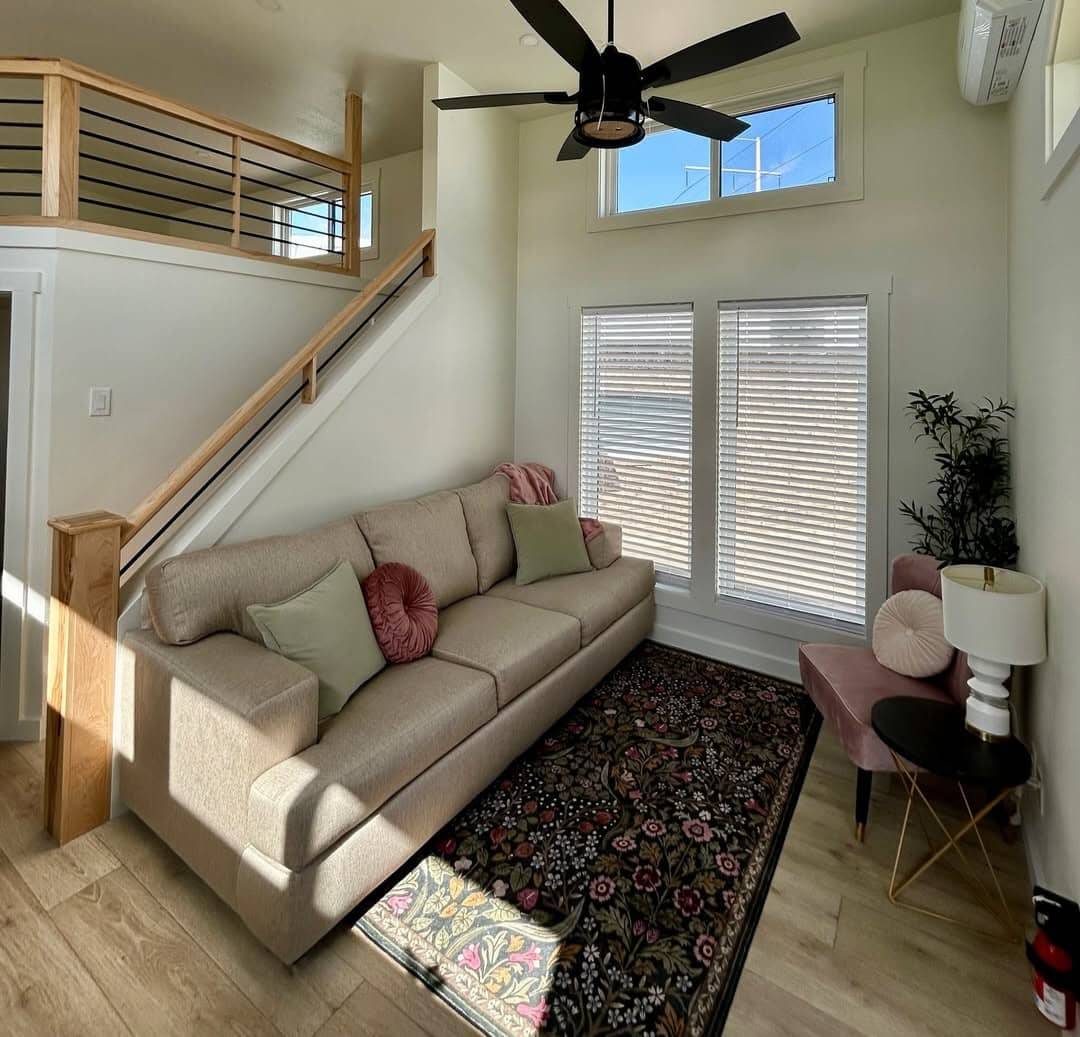 A cozy living room with a beige sofa, green and red pillows, floral rug, and a pink chair. Sunlight streams through tall windows, creating a warm atmosphere.
